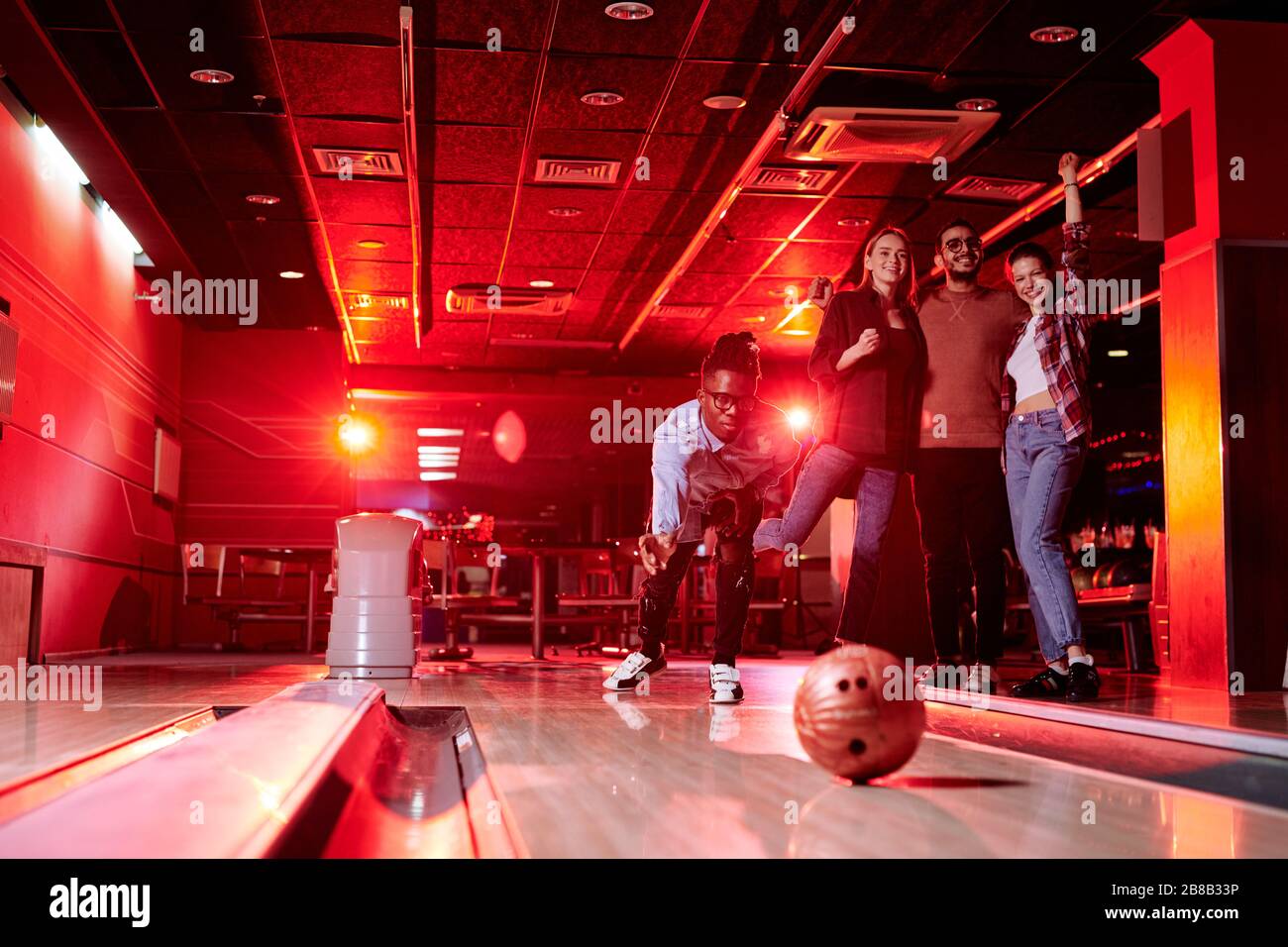 African guy bending by bowling alley while throwing ball on background of his friends with raised arms expressing gladness Stock Photo