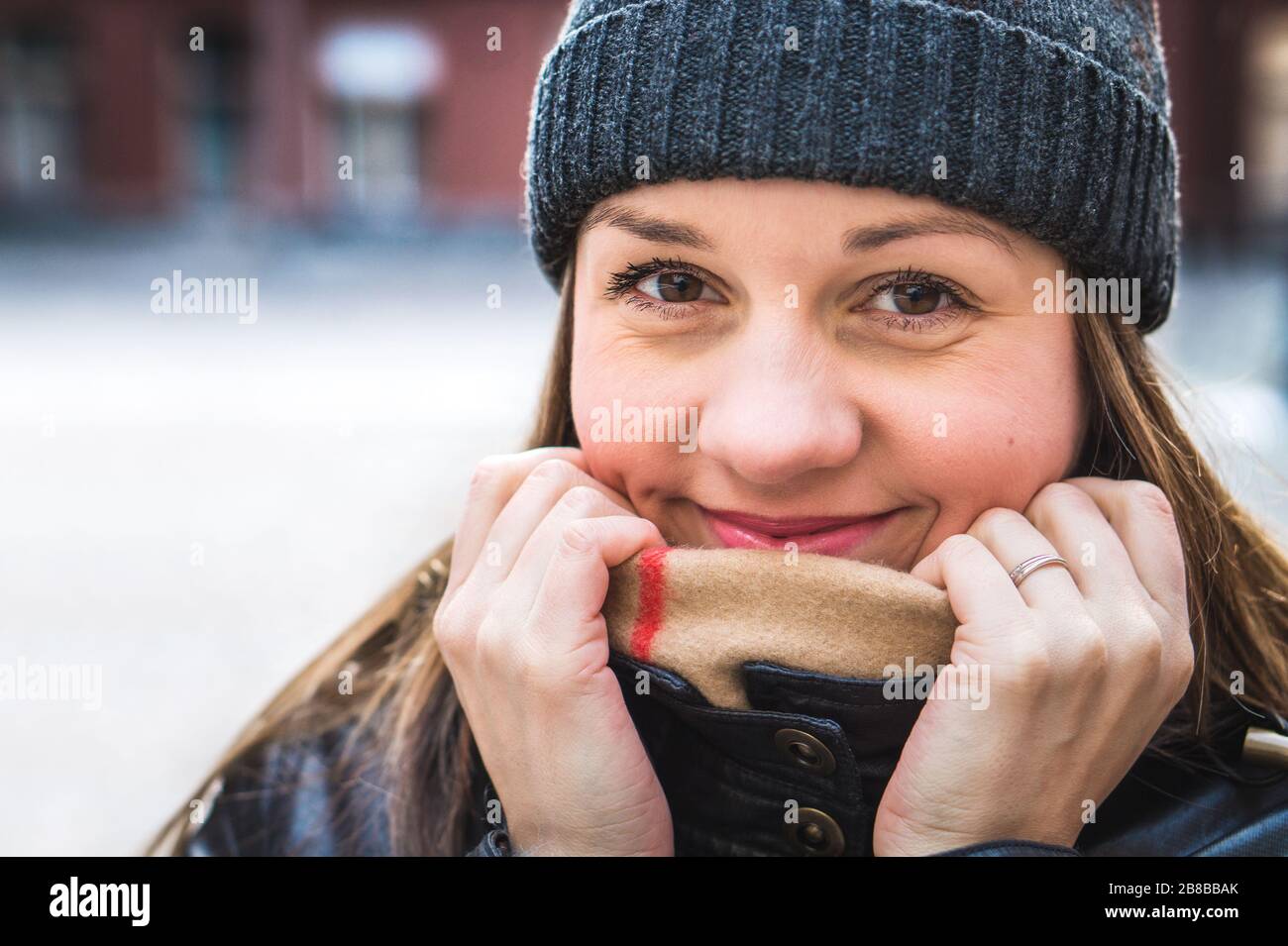 Cute woman wearing a beanie in winter. Happy and smiling person hiding behind scarf and holding collar with hands. Outdoor lifestyle portrait in city. Stock Photo