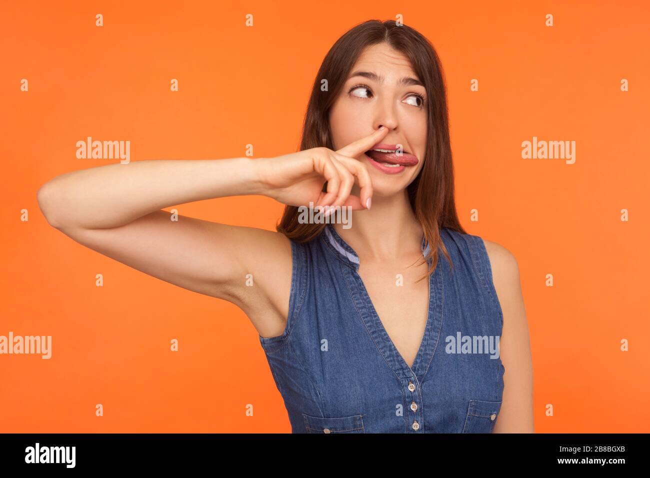 Funny comical brunette woman in denim dress picking nose and showing tongue with humorous stupid expression, uncultured habit, bad manners concept. in Stock Photo