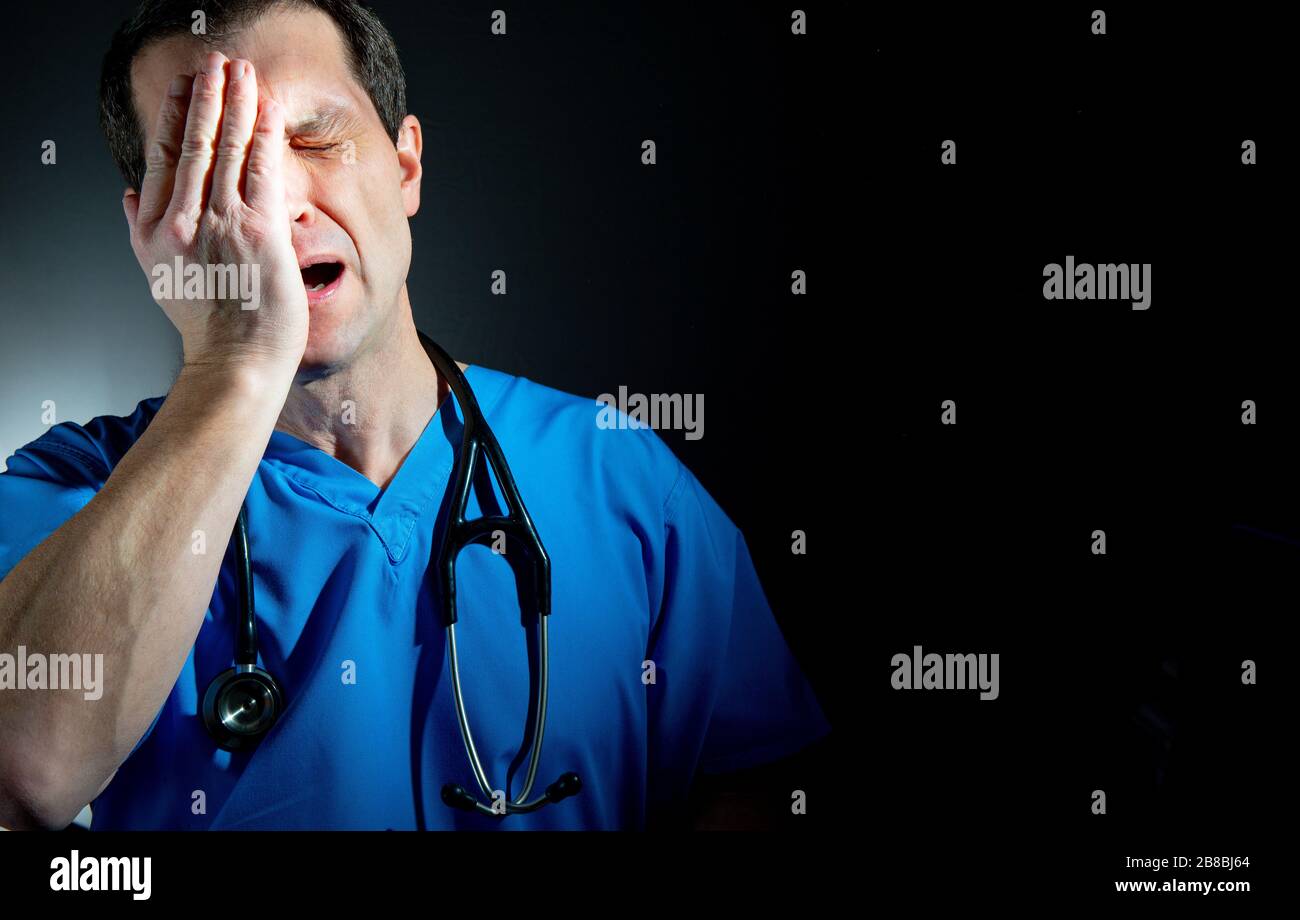 Upset / tearful doctor with hand on face, wearing blue scrubs, surgical face mask and stethoscope, against a dark background. Stock Photo
