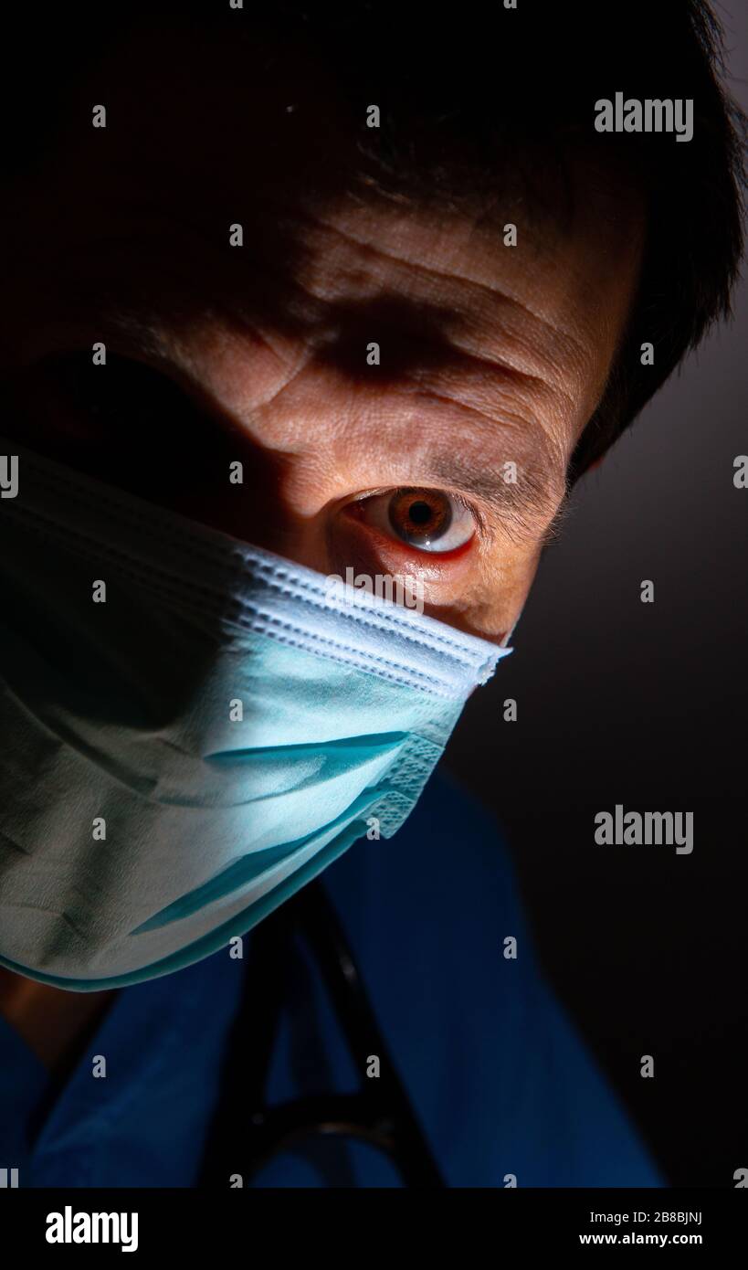 Close up of a concerned doctor with half face in shadow and wearing blue scrubs, with surgical face mask and stethoscope, against a dark background. Stock Photo