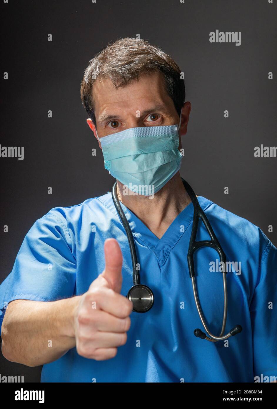 Doctor doing a thumbs up hand gesture, wearing blue scrubs, surgical face mask and stethoscope, against a dark background. Stock Photo