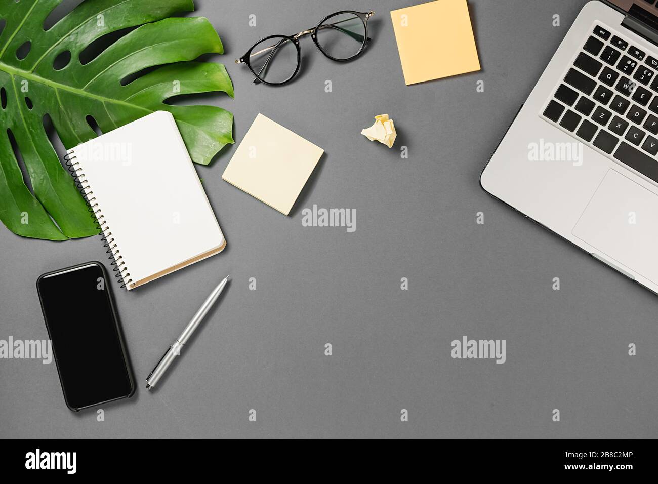 Workspace with modern smartphone, laptop, pen, monstera leaves, eyeglasses and notebook on grey background minimal style. Flat lay, top view office ta Stock Photo