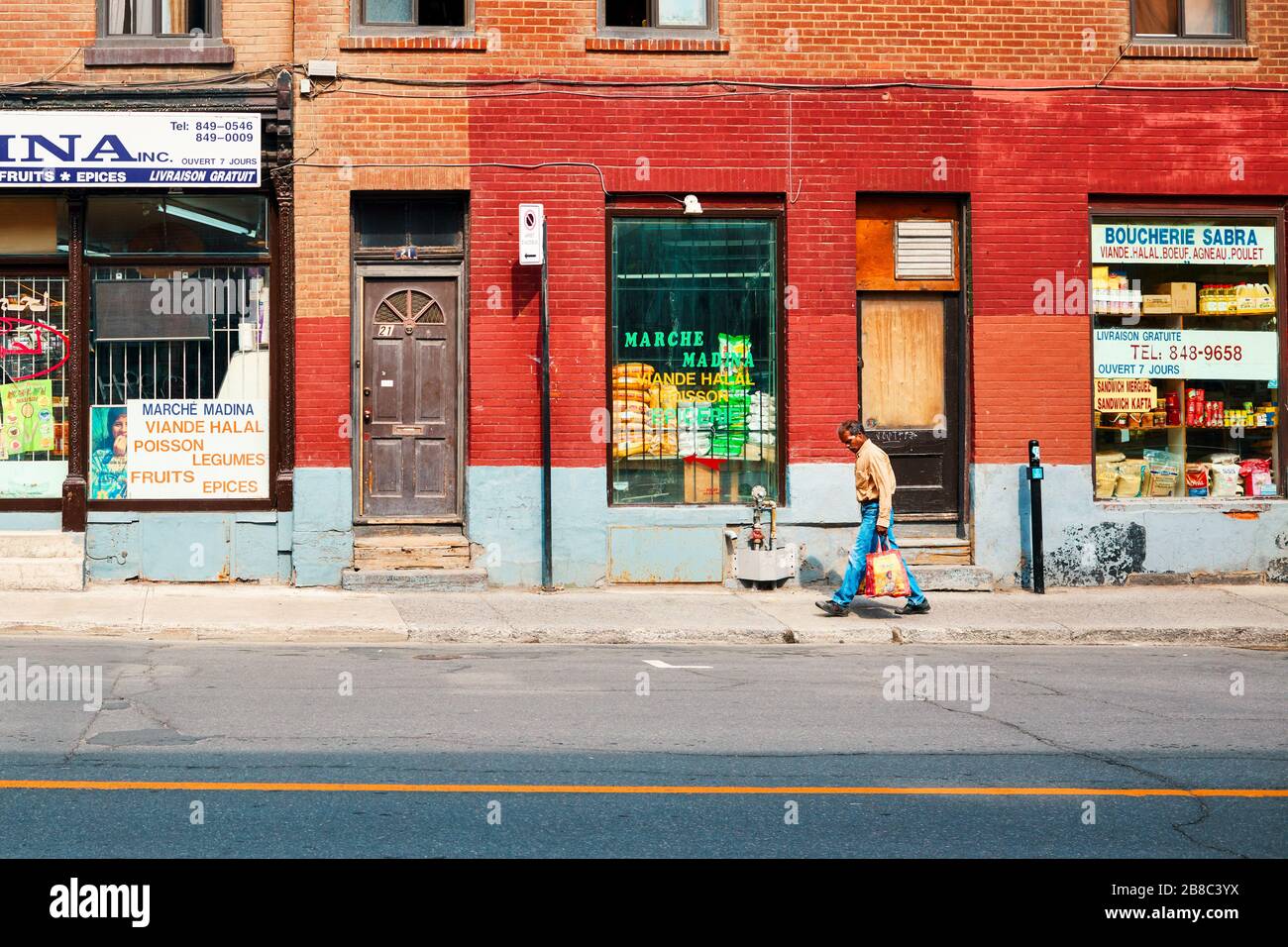 June, 2018 - Montreal, Canada: Indian or Pakistani man carrying shopping bags walks on Ontario street with historical buildings and shops in Montreal, Stock Photo