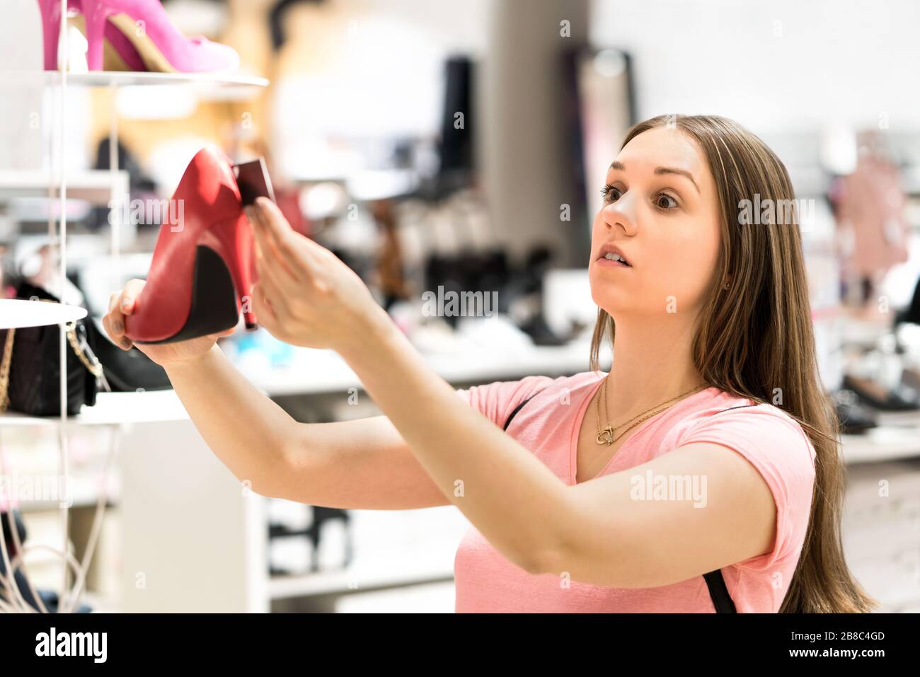 Shocked woman looking at price tag of too expensive shoes in fashion store while shopping. Unhappy customer holding high heels. Stock Photo