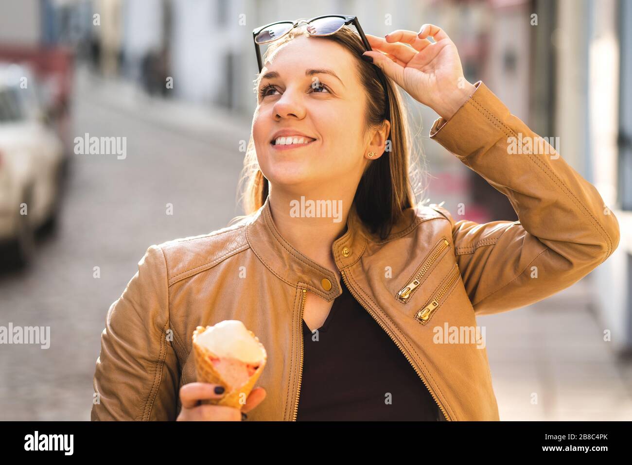 Happy woman eating ice cream and lifting sunglasses and looking up. Tourist in old town with sweet dessert during travel. Positive lifestyle at sunset. Stock Photo