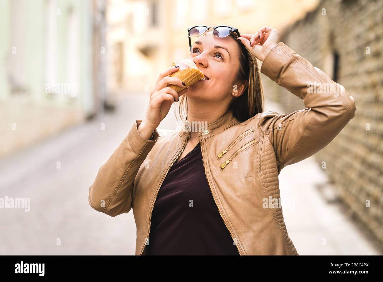 Woman lifting sunglasses and looking up while eating ice cream in city street. Tourist eating sweet dessert during travel. Stock Photo