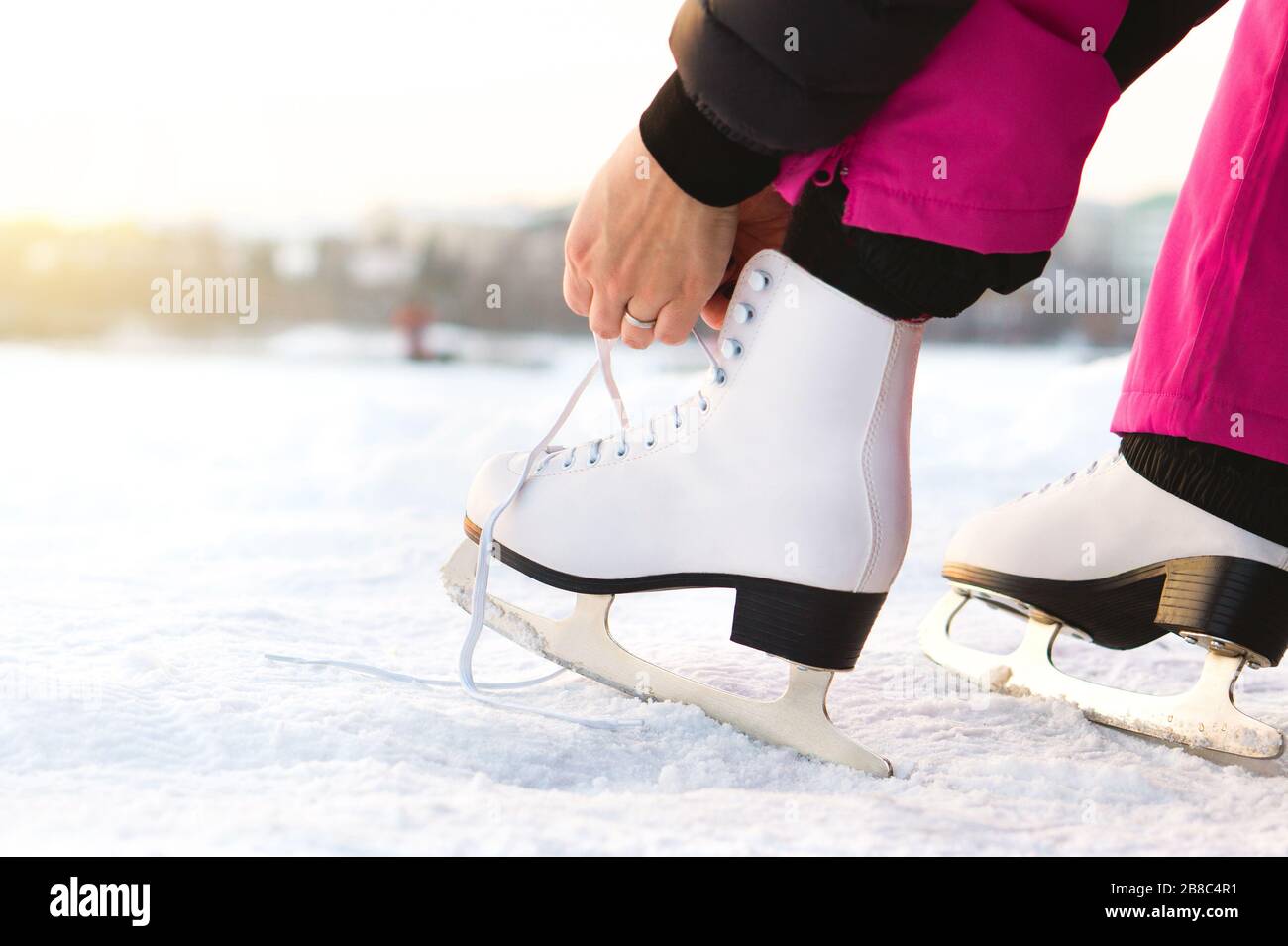 Woman tying ice skates laces by a lake or pond. Lacing iceskates. Skater about to exercise on an outdoor track or rink. Sunny winter weather. Stock Photo