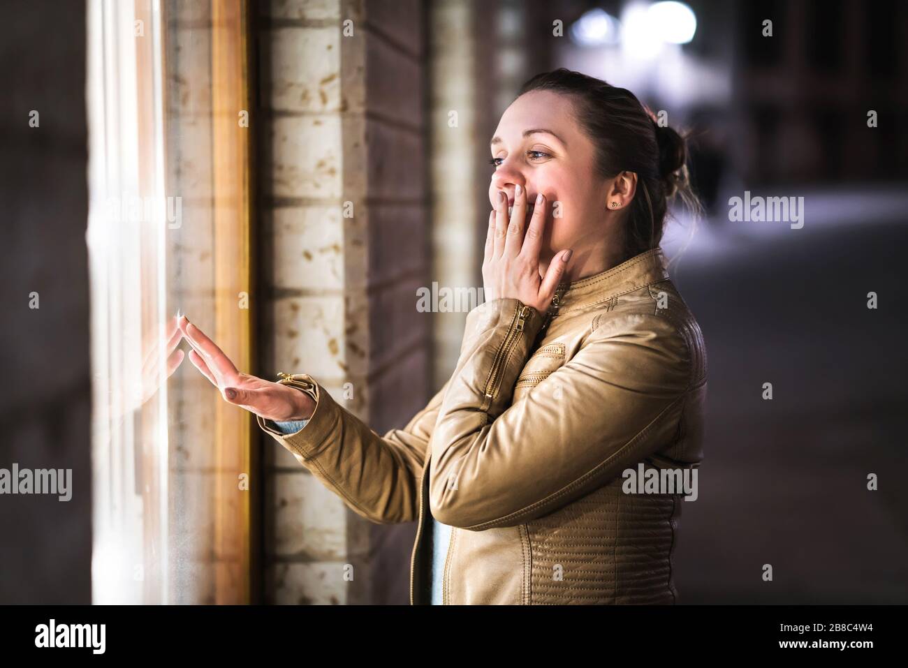 Shopaholic window shopping. Woman admiring and dreaming about new shoes, bag or clothes. Smiling lady covering mouth and touching fashion store glass. Stock Photo