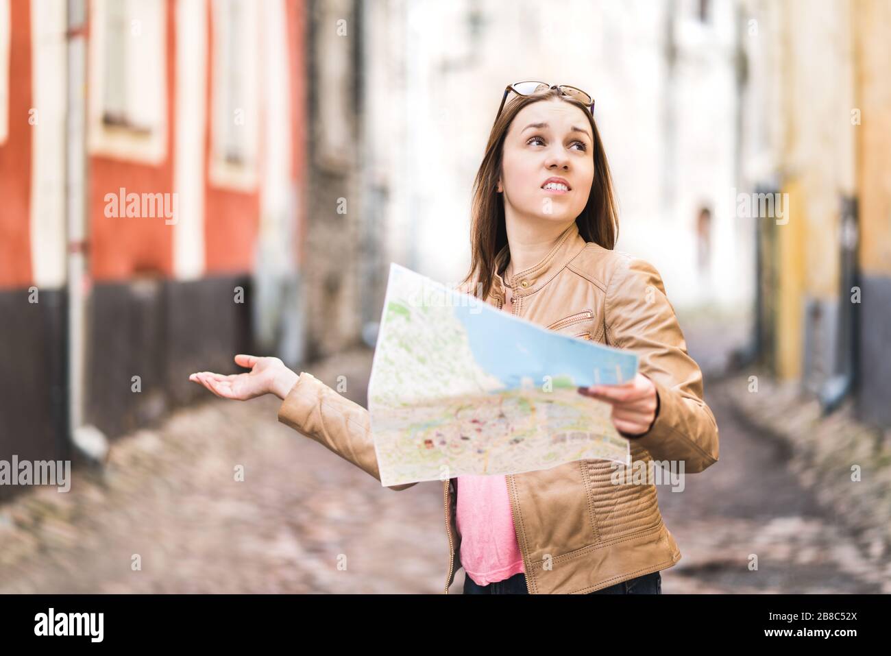 Woman lost in the city. Confused traveler holding map and spreading hands in old town. Disappointed and worried tourist having problem. Stock Photo