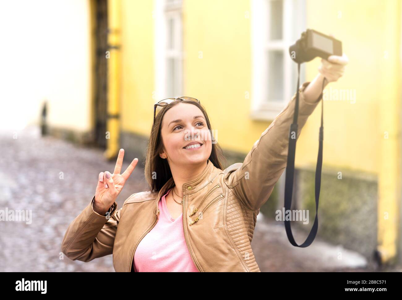 Vlogger filming video blog in city. Young smiling happy woman making vlog or taking selfie with camera. Showing v sign hand gesture. Stock Photo