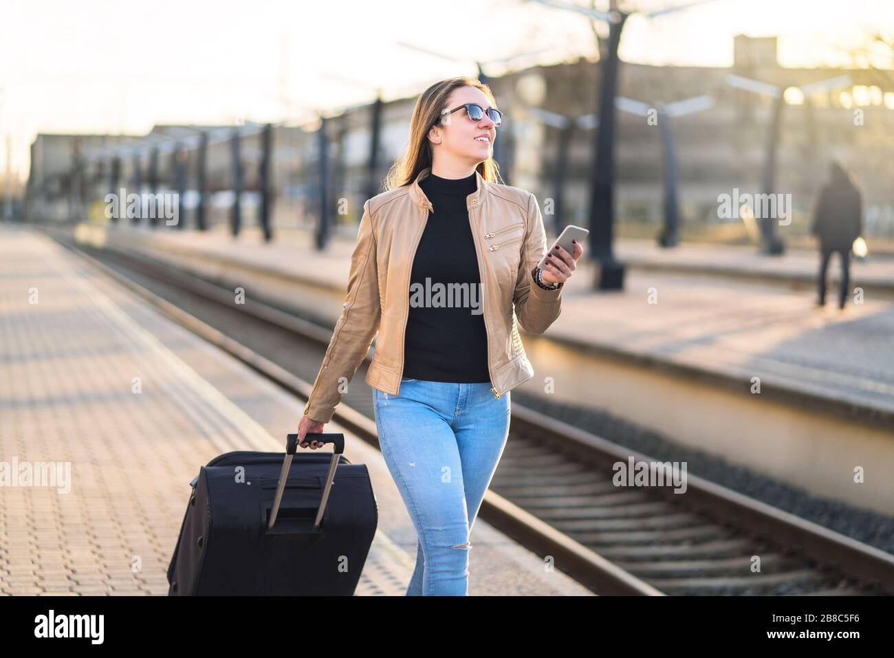 Confident business woman pulling suitcase and holding mobile phone in train station. Smiling lady walking in platform and using smartphone. Stock Photo