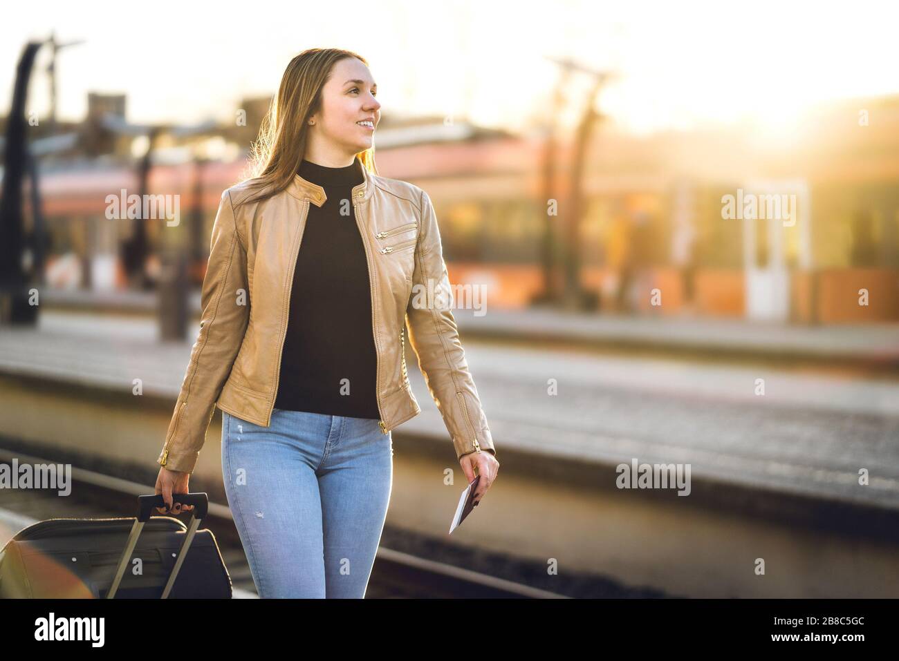 Happy woman pulling suitcase in train station. Smiling lady with baggage and luggage arrived to destination. Tourist on vacation. Stock Photo