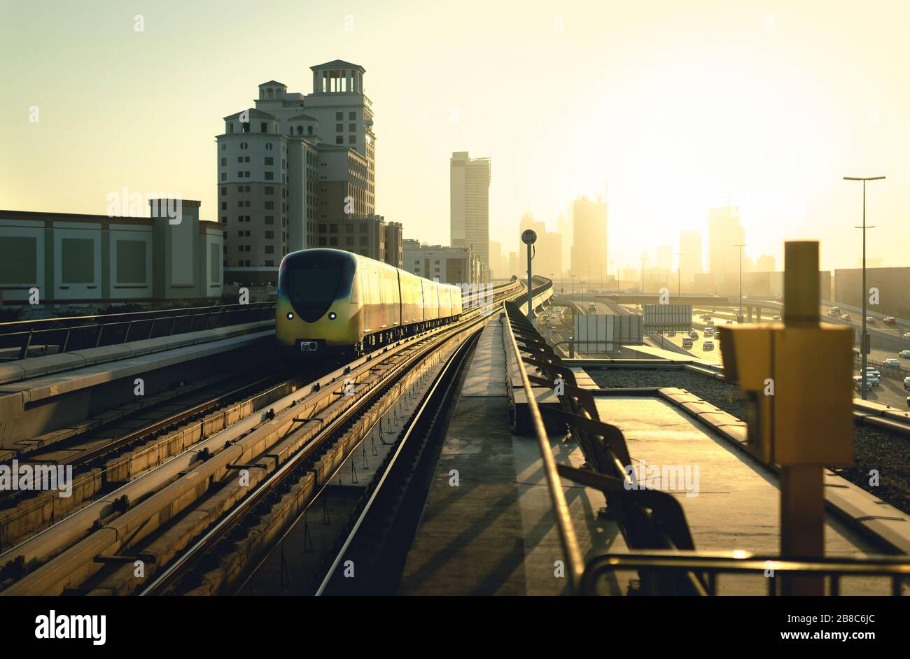 Dubai metro at sunset. Modern subway, car traffic on highway and business buildings. City downtown skyline and railway at golden hour dusk. Stock Photo