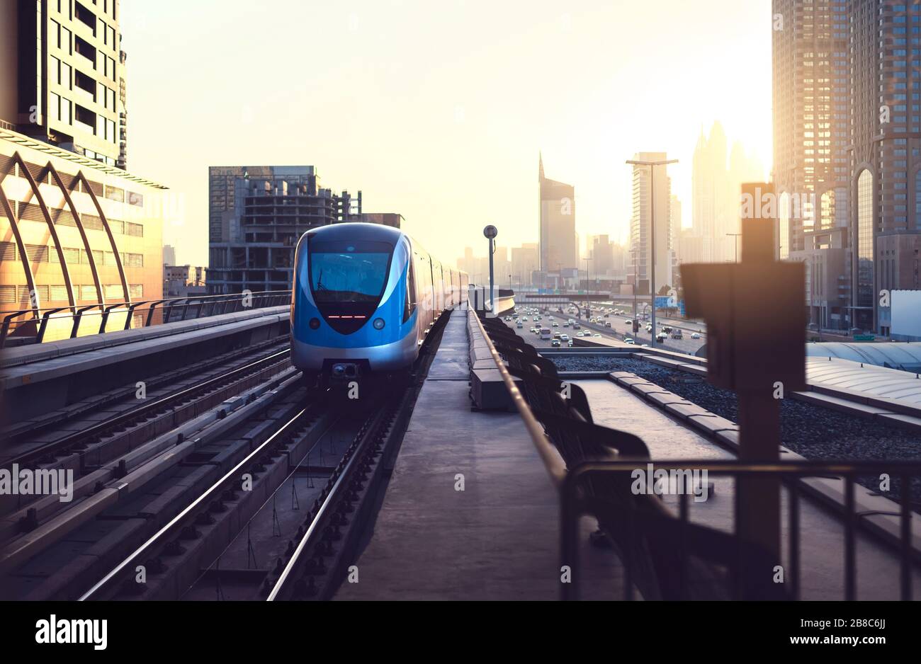 Subway train at sunset in modern city. Dubai metro. Downtown skyline with sundown. Skyscraper buildings and car traffic in the highway. Stock Photo