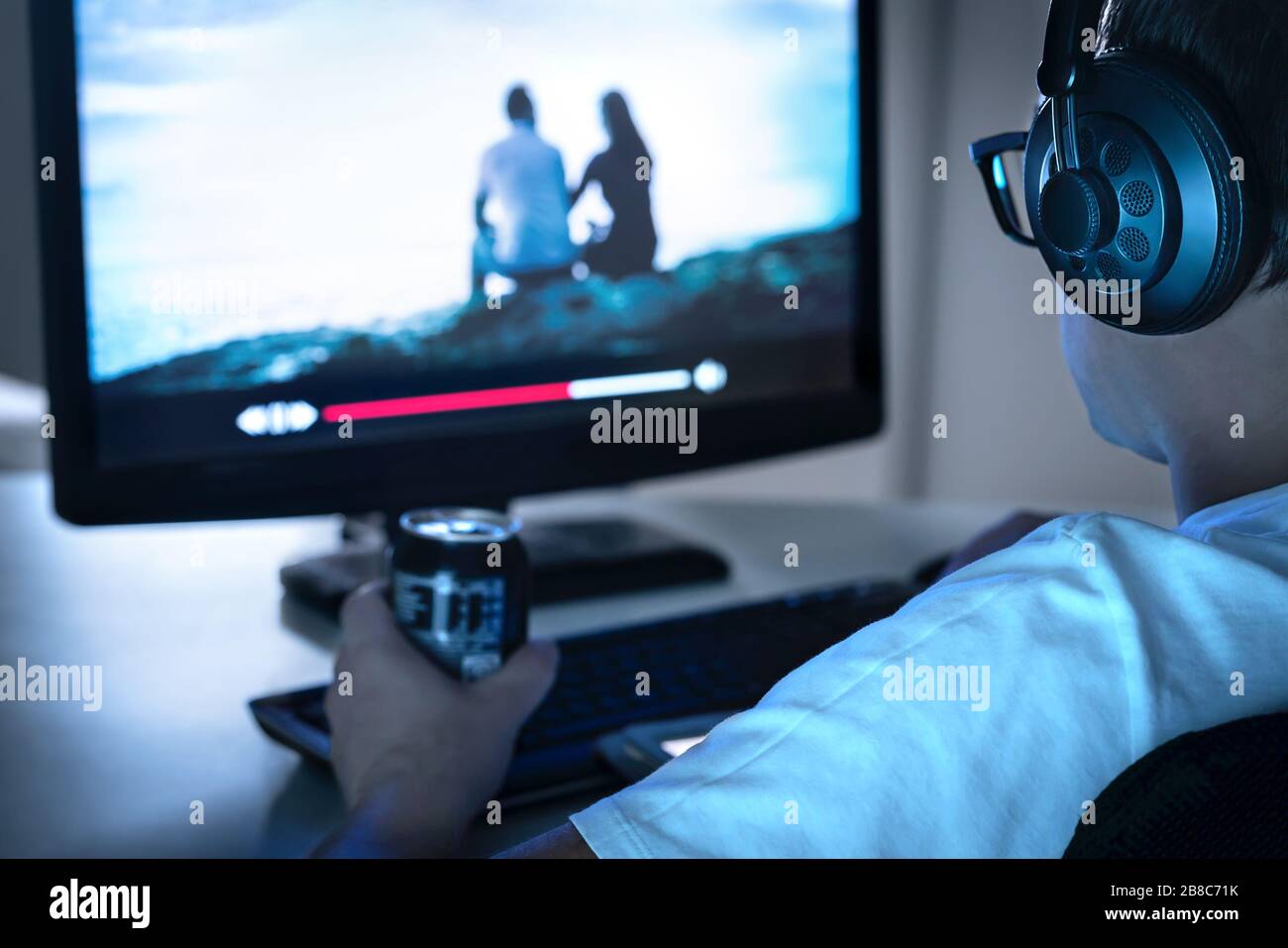 Guy watching movie or series online from streaming service and drinking can of soda. Internet on demand video player in computer. Stock Photo
