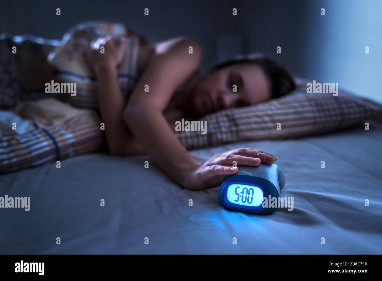 Tired woman waking up for work or school early in the morning. Grumpy lady pushing snooze button or turning off alarm clock with hand. Stock Photo