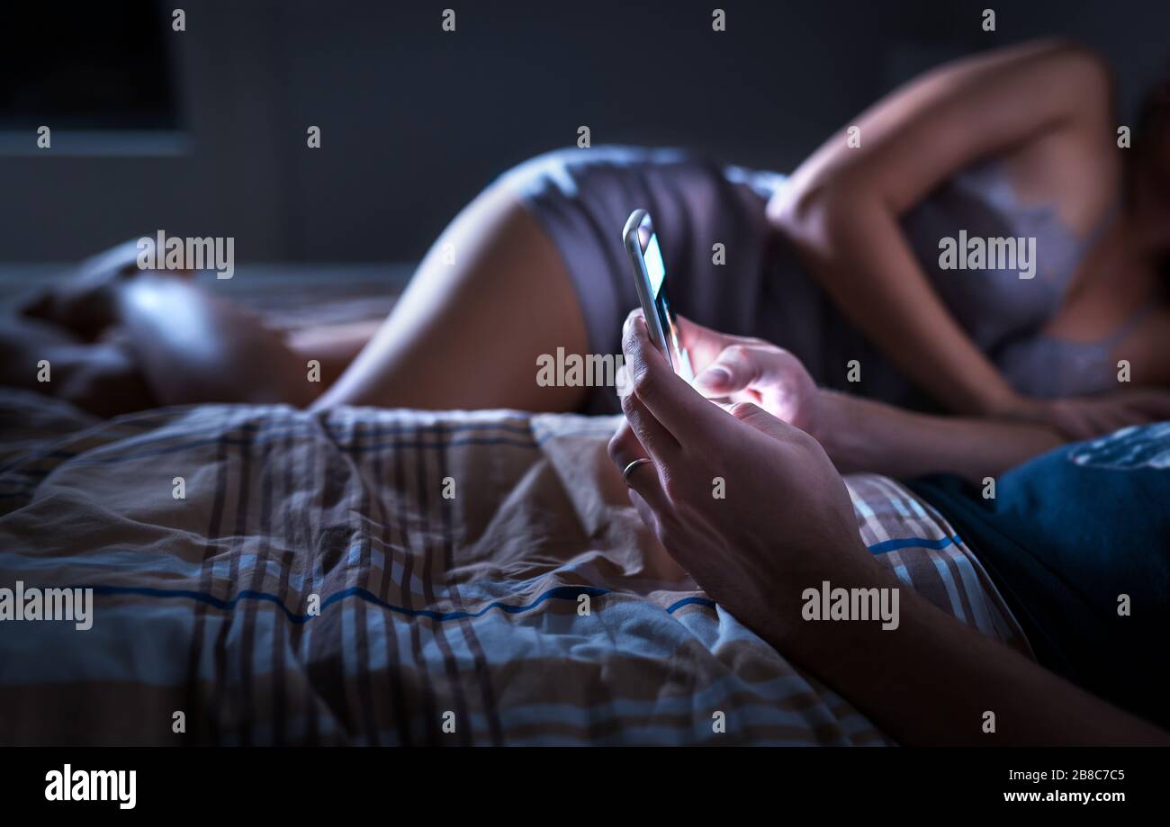 Man using smartphone and ignoring sexy woman in bed at night. Looking at mobile phone. Husband giving no attention to wife. Disregarding marriage. Stock Photo