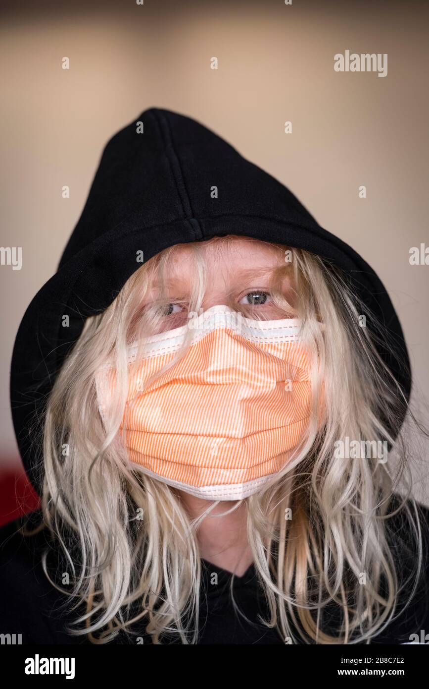 12-year old boy with long blond hair is wearing an infection protection face mask and black hoodie. Caucasian ethnicity, face covered by hair and face Stock Photo
