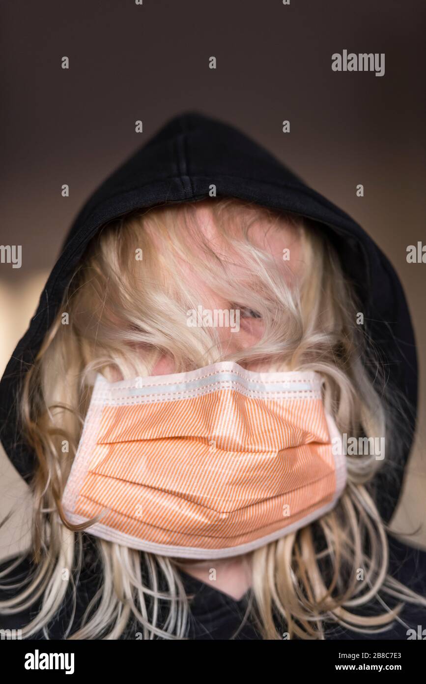 12-year old boy with long blond hair is wearing a slipped infection protection face mask and black hoodie. Caucasian ethnicity, face covered by hair a Stock Photo