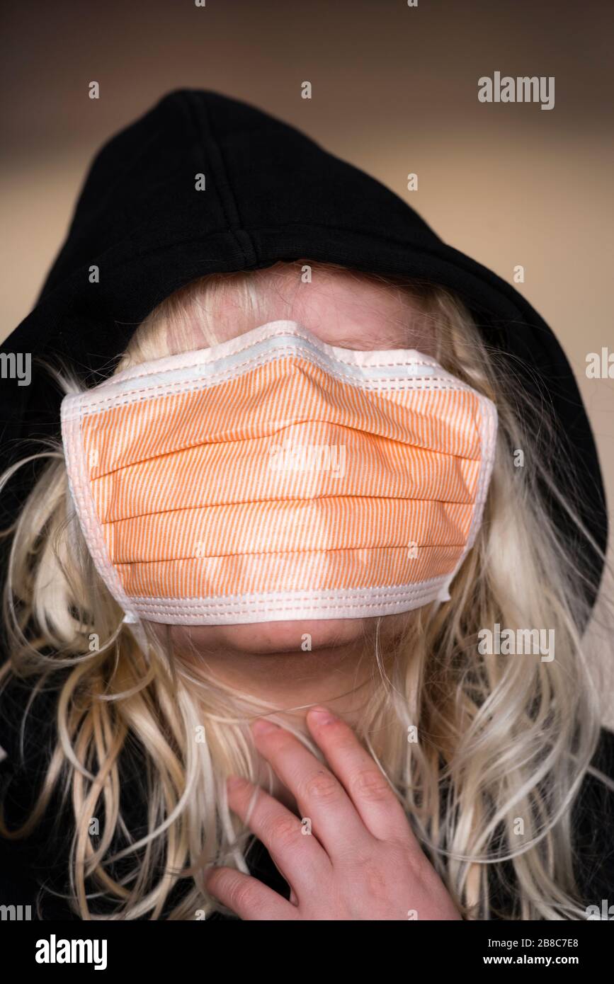 12-year old boy with long blond hair is wearing an infection protection face mask and black hoodie. Caucasian ethnicity, face covered completely by fa Stock Photo