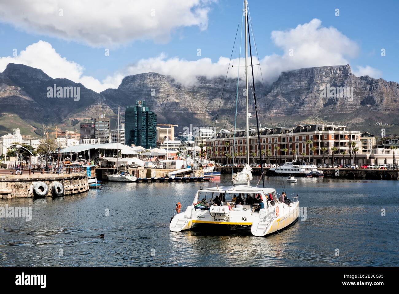 A catamaran loaded with tourists sails towards the Cape Grace Hotel on the V & A Waterfront while Table Mountain looks down at the city from behind Stock Photo