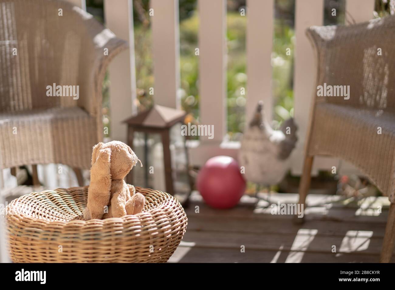 Stuffed toy easter bunny sitting on a straw chair on the veranda, looking to the sun. High angle view, close up, concept. Stock Photo