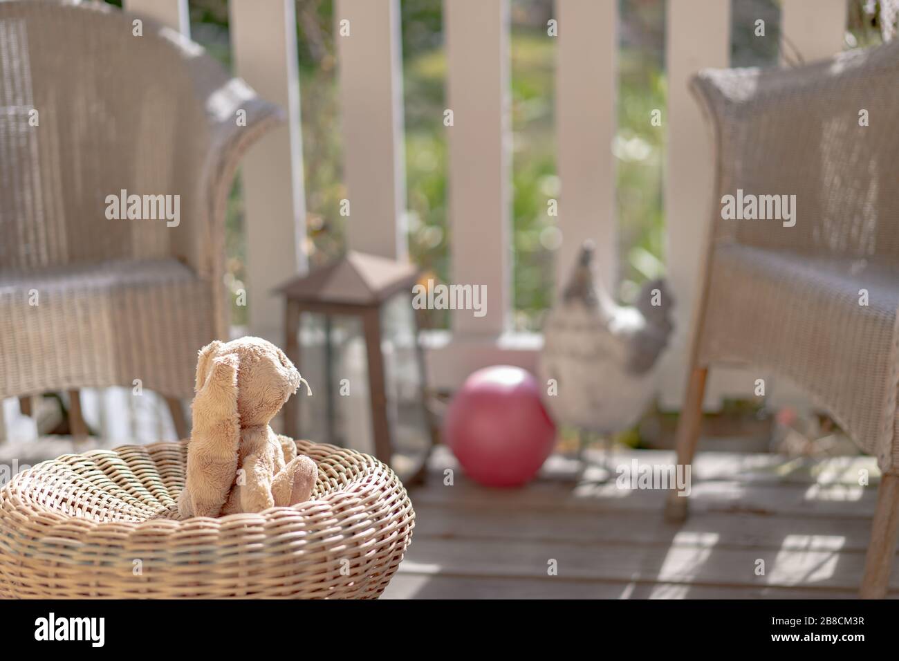 Stuffed toy easter bunny sitting on a straw chair on the veranda, looking to the sun. High angle view, close up, concept. Stock Photo