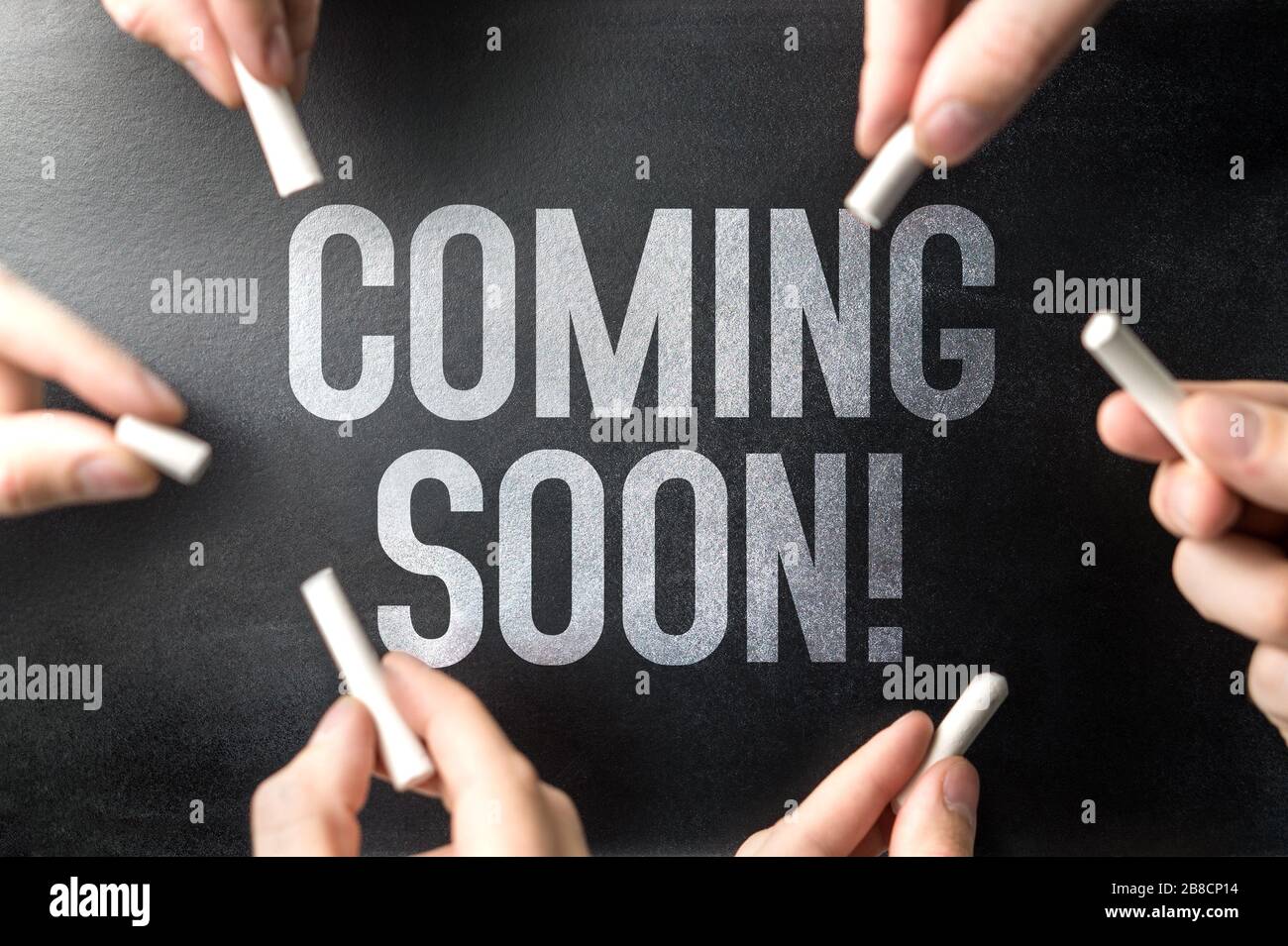 Coming soon text written on chalkboard sign and blackboard banner by many hands. Website or web page under construction or new announcement. Stock Photo