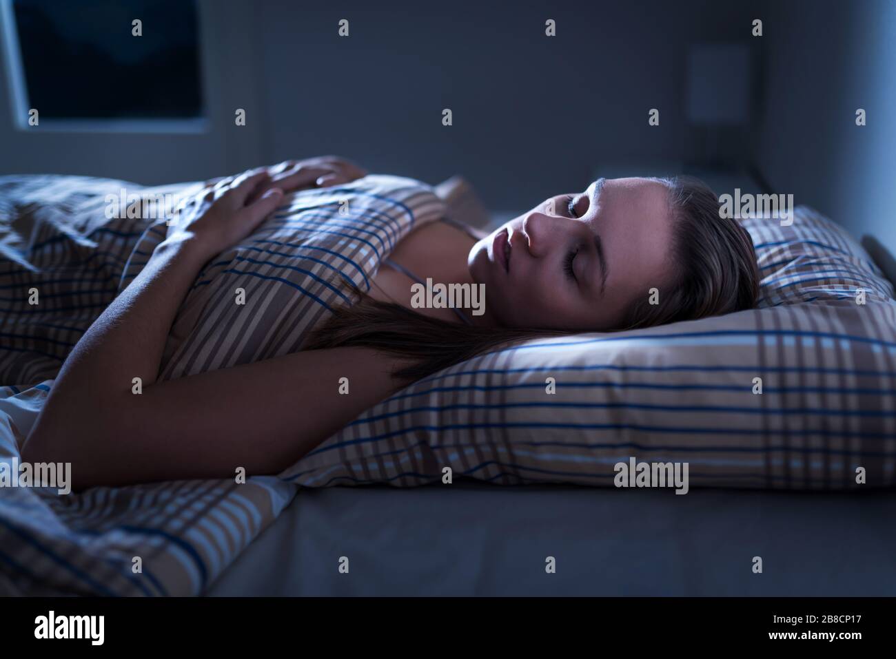 Calm and peaceful woman sleeping in bed in dark bedroom. Lady asleep at home in the middle of the night. Pillow, blanket and moonlight. Stock Photo