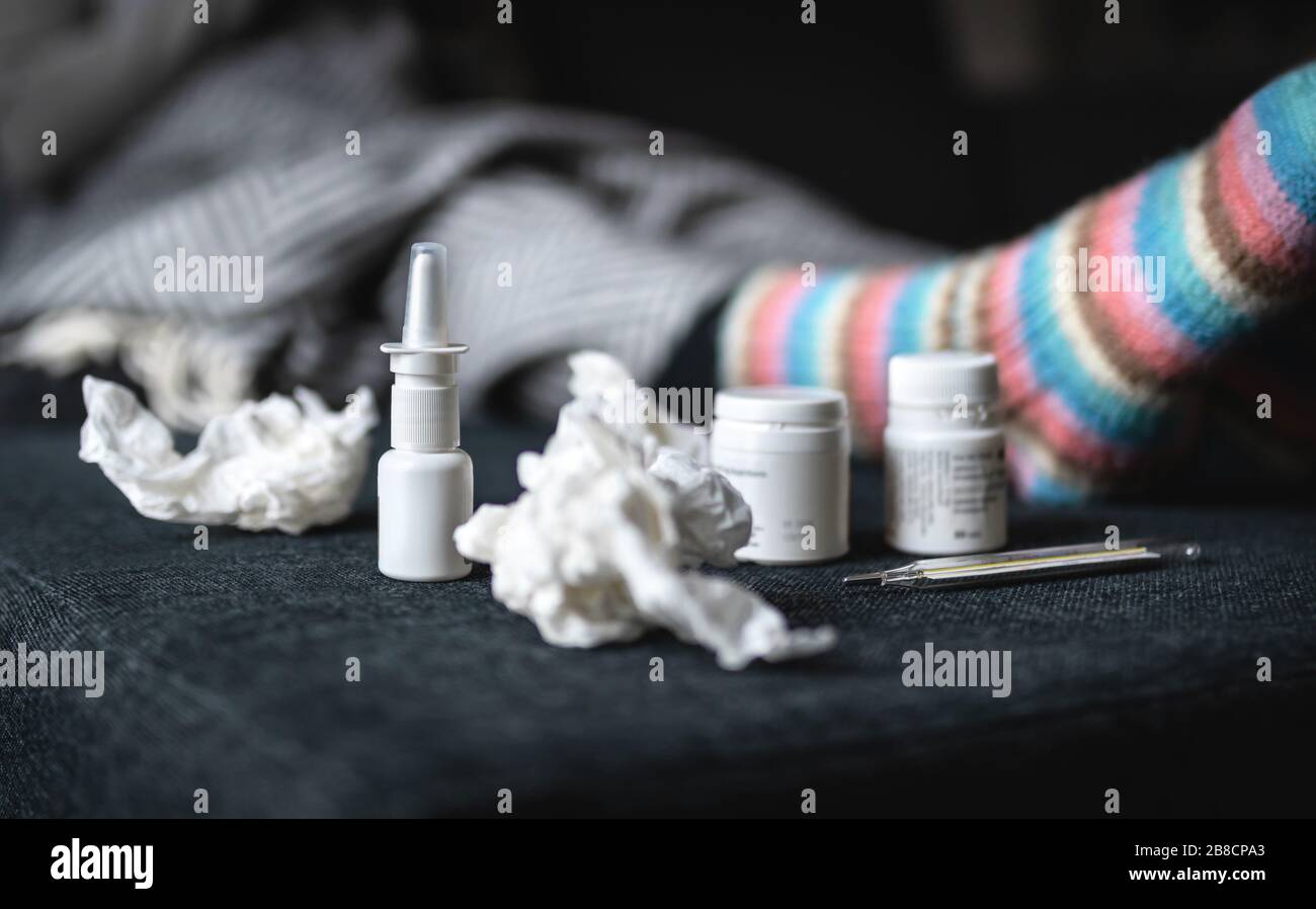Flu and cold medicine. Sick woman with nasal spray, warm woolen socks, tissues and thermometer resting on couch at home. Ill person lying on sofa. Stock Photo