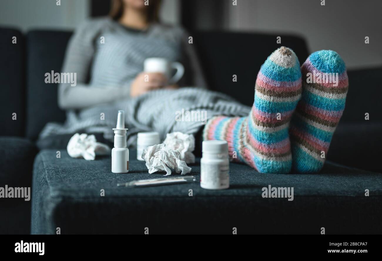 Sick woman resting on couch holding hot cup of tea. Ill person with flu, cold, fever or virus sitting on sofa at home in winter. Medicine, thermometer. Stock Photo