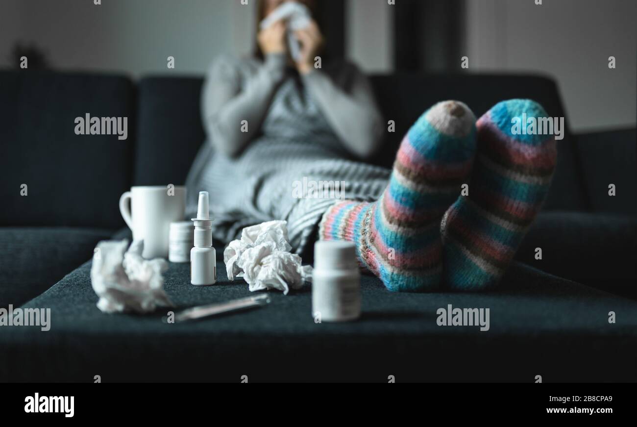 Woman sneezing and blowing nose with tissue and handkerchief. Sick and ill person with flu, cold medicine and woolen socks. Fever, virus or infection. Stock Photo