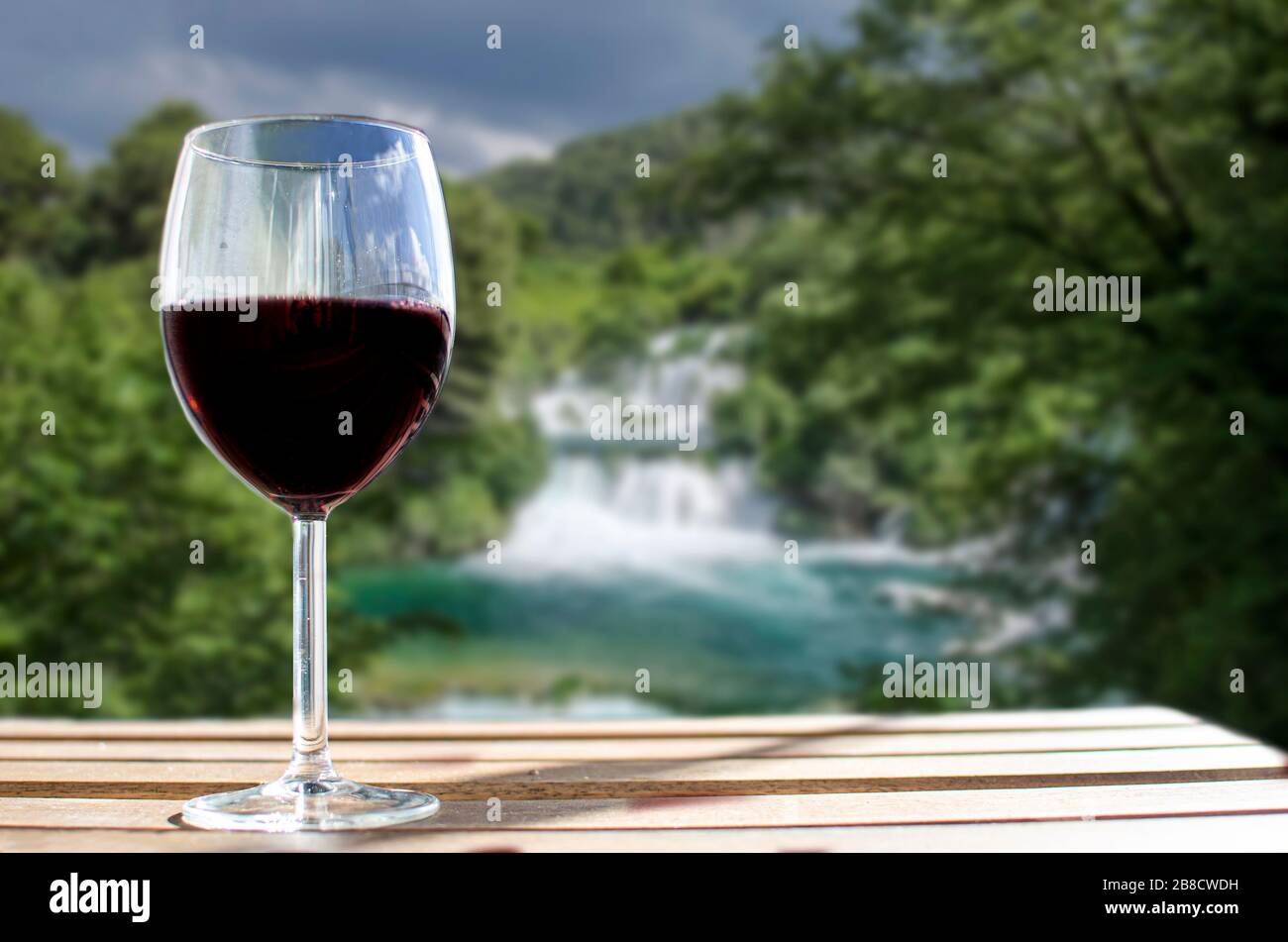 Glass of wine on wooden table with big waterfall blur background in Croatia. Sunny view of glass of red wine overlooking park, Croatia Stock Photo