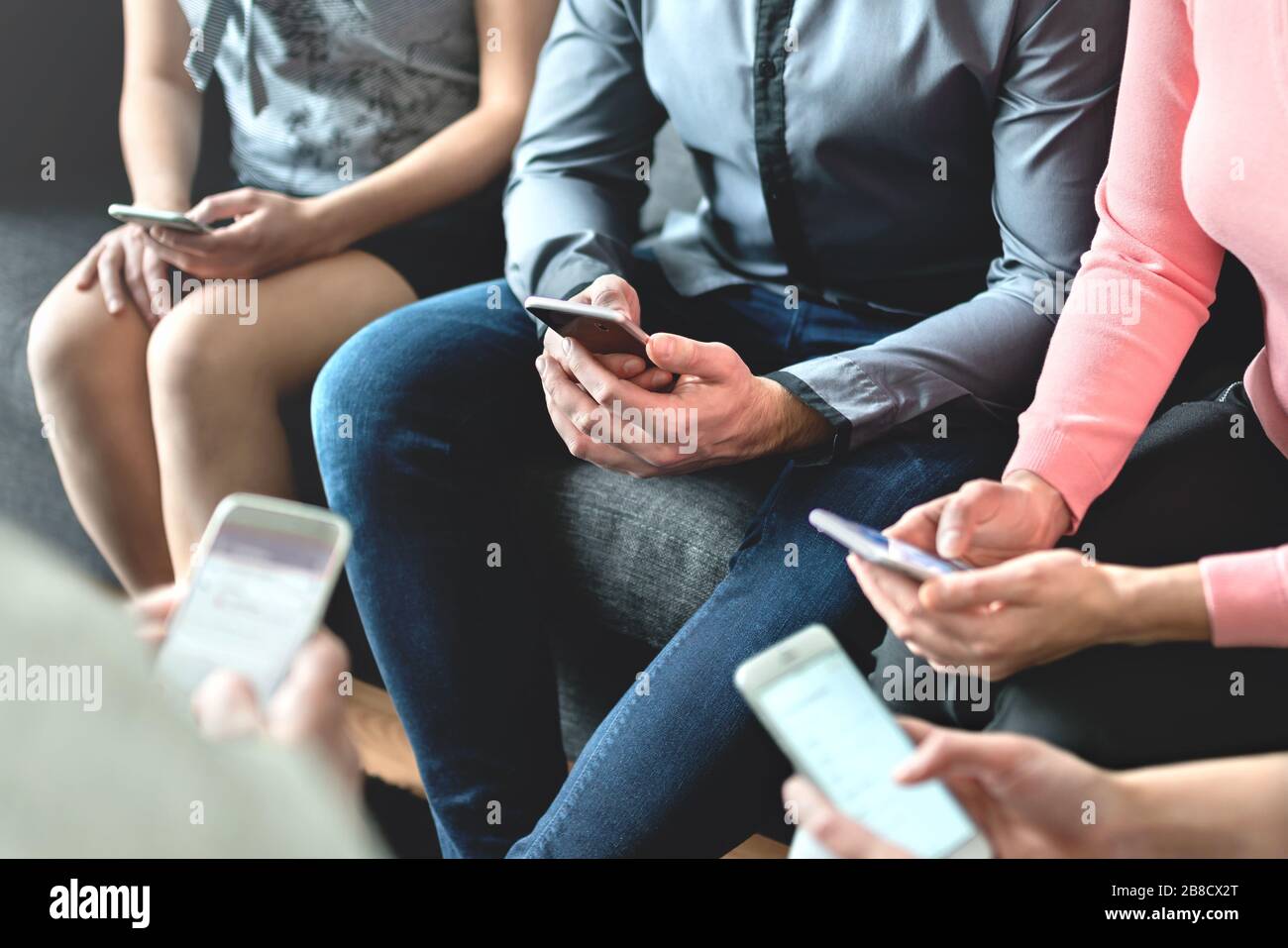 Group of millennial business people using mobile phones and sitting on couch. Networking and teamwork. Working on project with wireless technology. Stock Photo