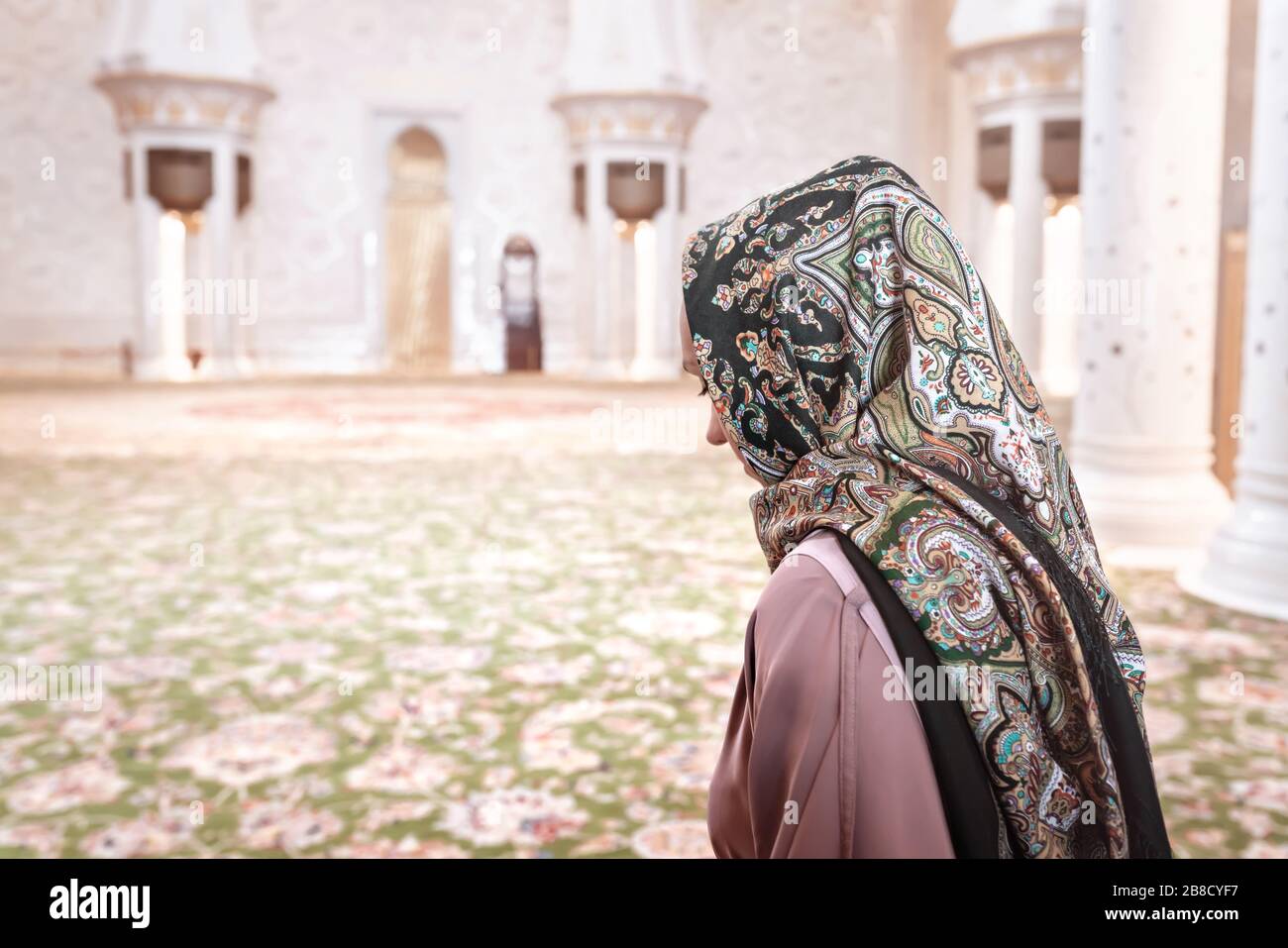 Woman in Muslim prayer room in mosque. Young lady wearing headscarf. Traditional carpet and Arab architecture. Islam religion and tourism concept. Stock Photo