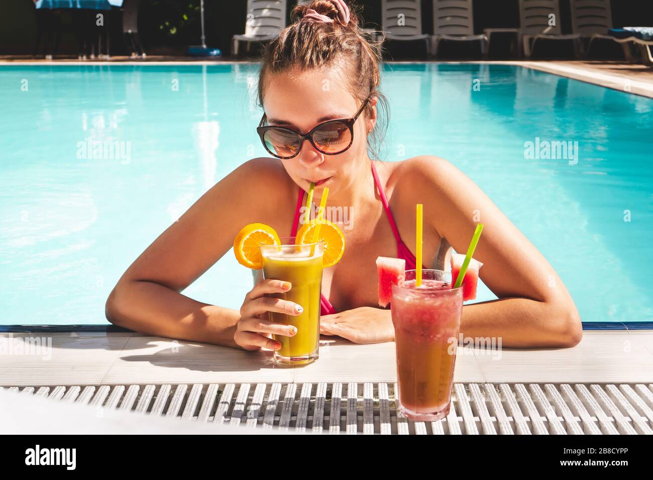 Woman drinking cocktail on the edge of swimming pool in an all inclusive hotel resort or luxury holiday villa. Tan lady in bikini in the poolside. Stock Photo