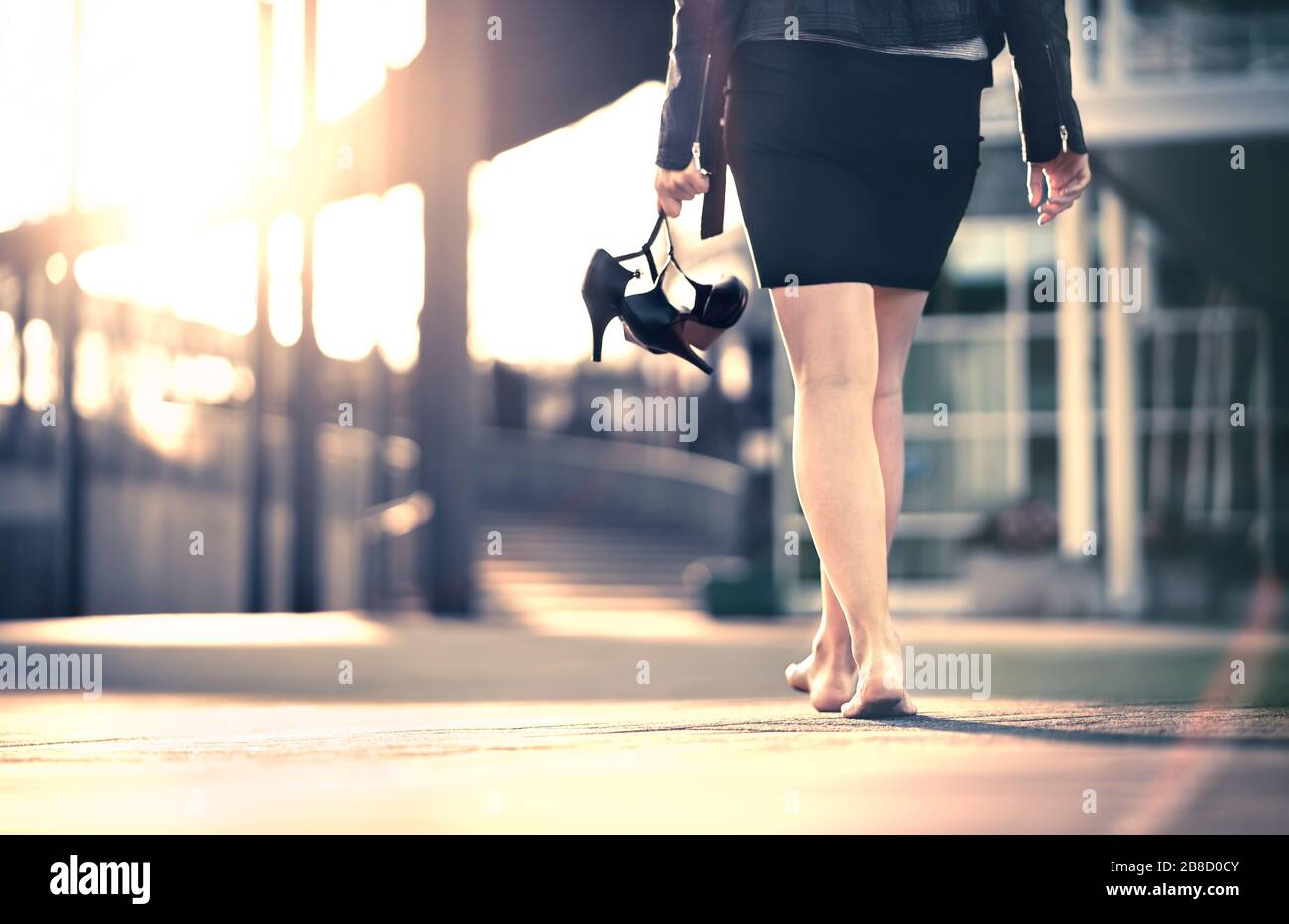 Woman holding high heels in hand and walking home from party barefoot. Businesswoman took off uncomfortable shoes. Lady with stilettos and miniskirt. Stock Photo