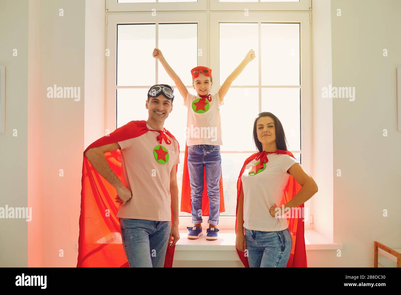 Happy cheerful family in costumes of super heroes laughing while standing against the background of a window in a room indoors. Stock Photo