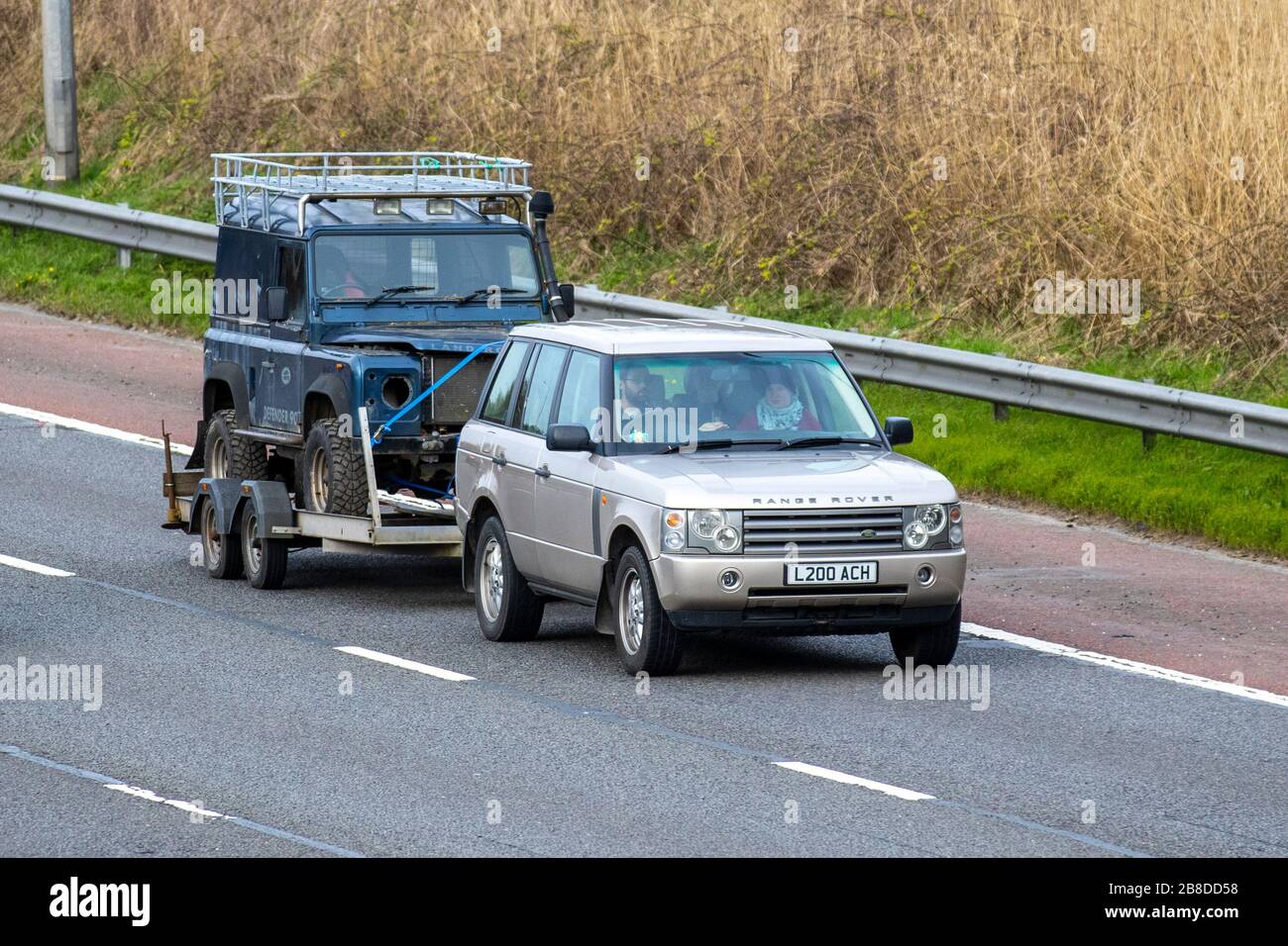 Range Rover towing decrepit dilapidated old Land Rover Defender. A restoration project, towed barn find; UK vehicular traffic, transport, moving vehicles, restoration vehicle, roads, towing motors, motoring trailer on the M6 motorway highway Stock Photo
