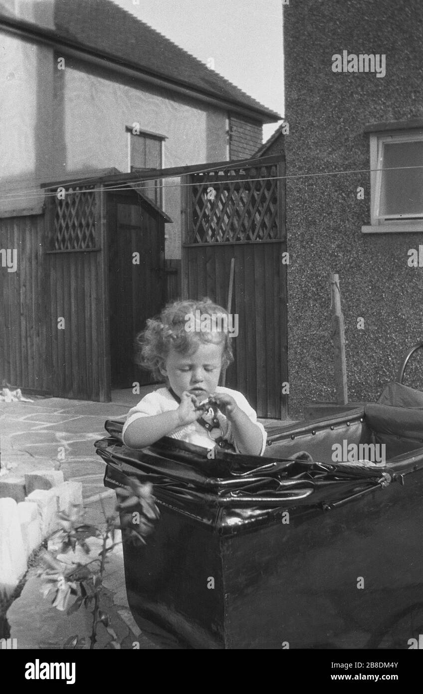 1953, historical, infant girl sitting inside an old leather lined pram, on a patio area outside at the back of a suburban pebble-dash house, England, UK. Stock Photo