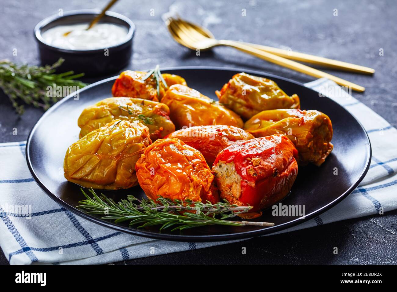 Classic stuffed bell peppers with rice, ground beef, and pork in tomato sauce, rosemary and thyme on a black plate with sour cream and gold cutlery, o Stock Photo