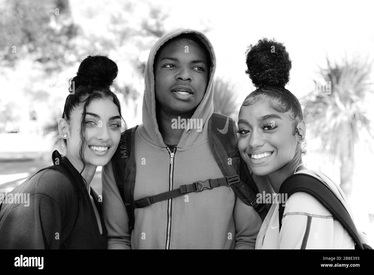 A college age African American man in hoodie between two female coeds  of color with trendy cool up-dos on campus enjoying student life, in black and Stock Photo