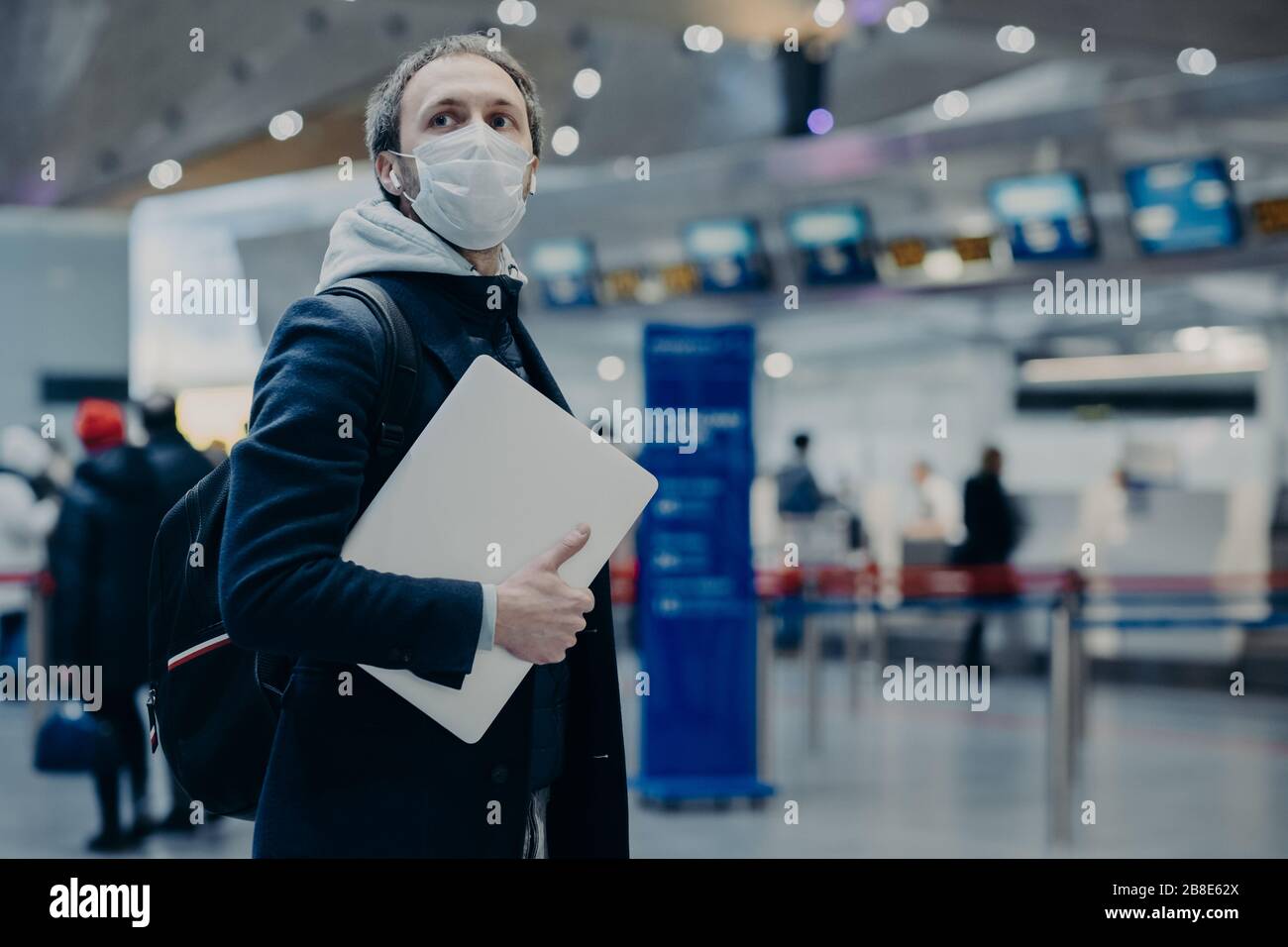 Man traveler wears protective disposable medical mask in airport, returns from abroad where coronavirus spreading, carries backpack, takes care of hea Stock Photo