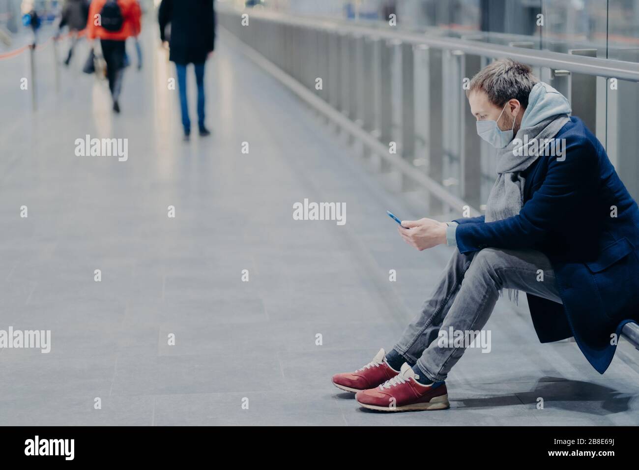 Horizontal shot of man holds modern mobile phone, being in mall during pandemic and coronavirus, wears protective medical mask, dressed casually, chec Stock Photo