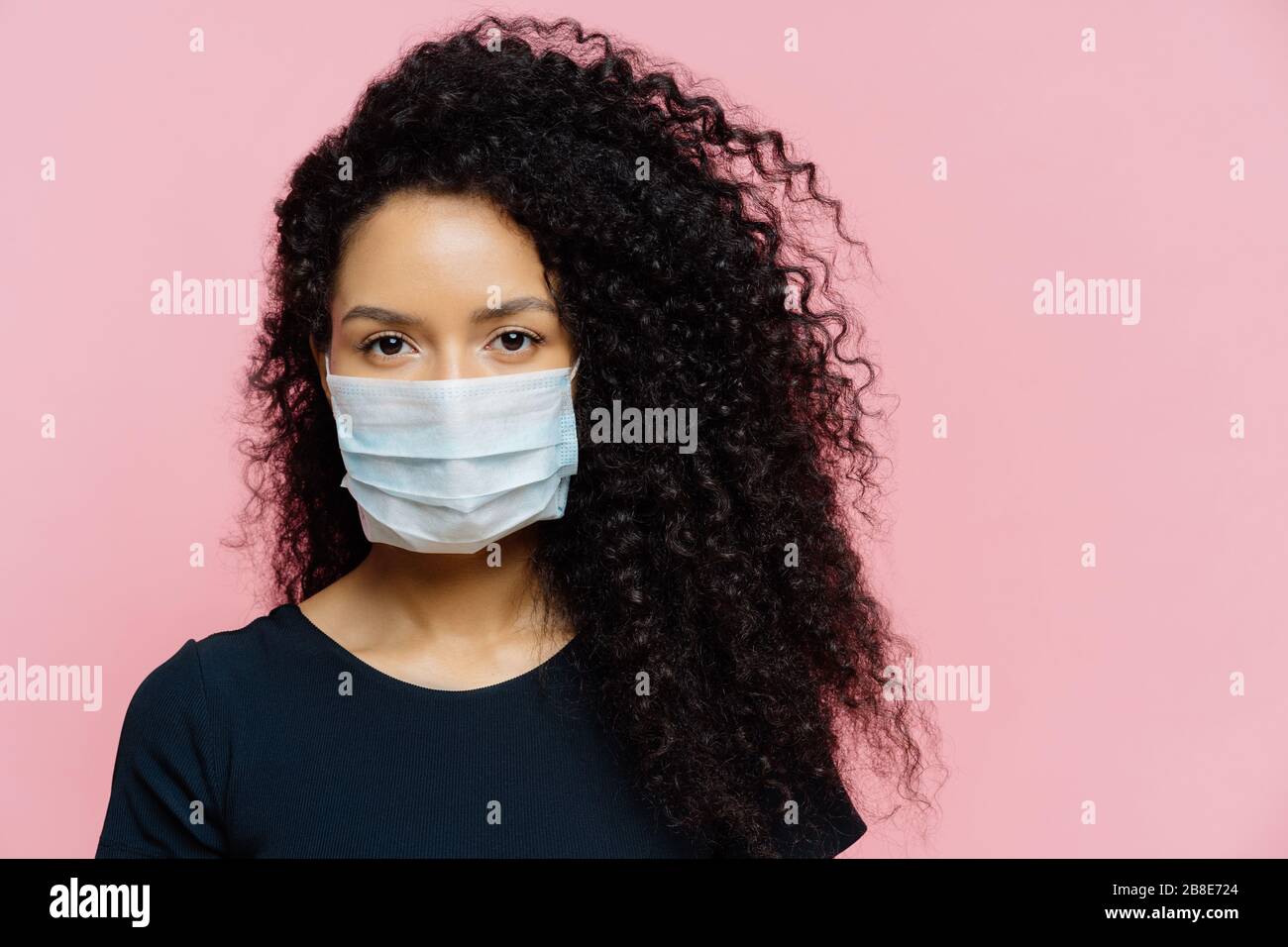 Serious dark skinned young woman being on self isolation at home, wears protective medical mask, being on quarantine at home, dressed casually, poses Stock Photo
