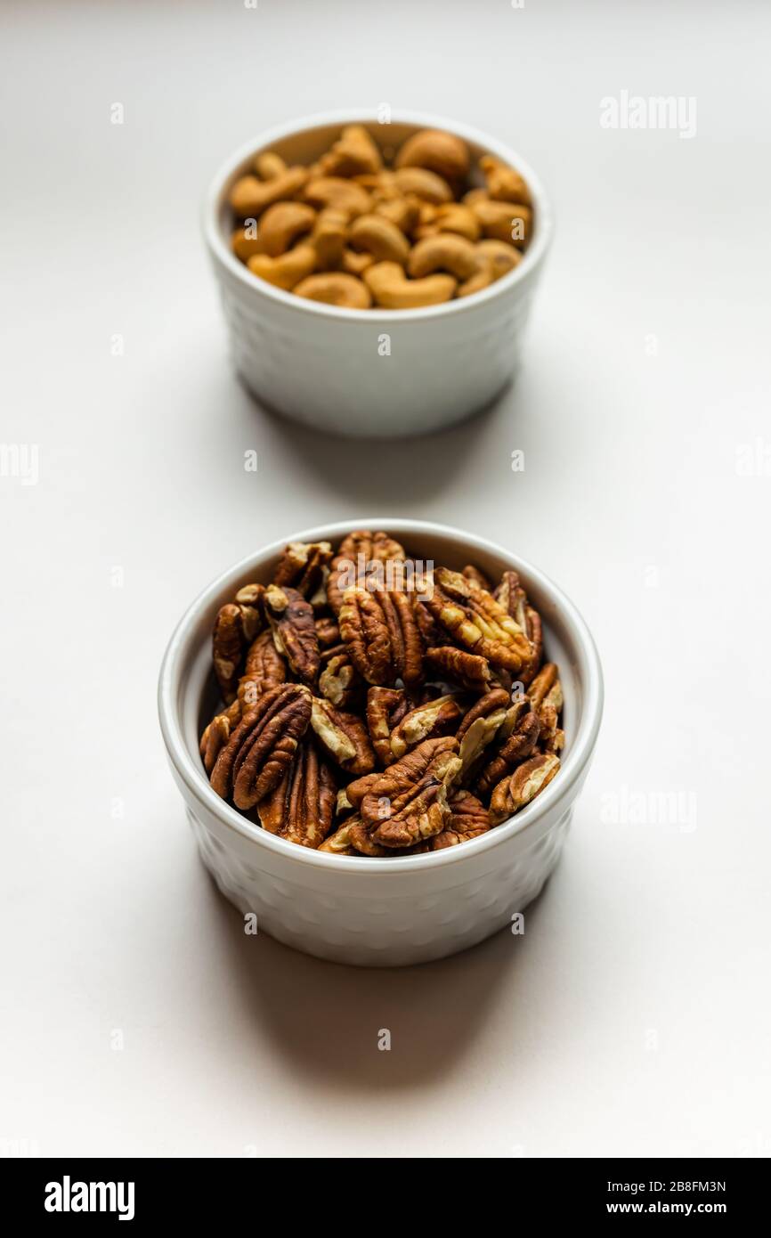 A small white bowl of delicious healthy salted pecan nuts in front of a white bowl of cashews isolated on a white background Stock Photo