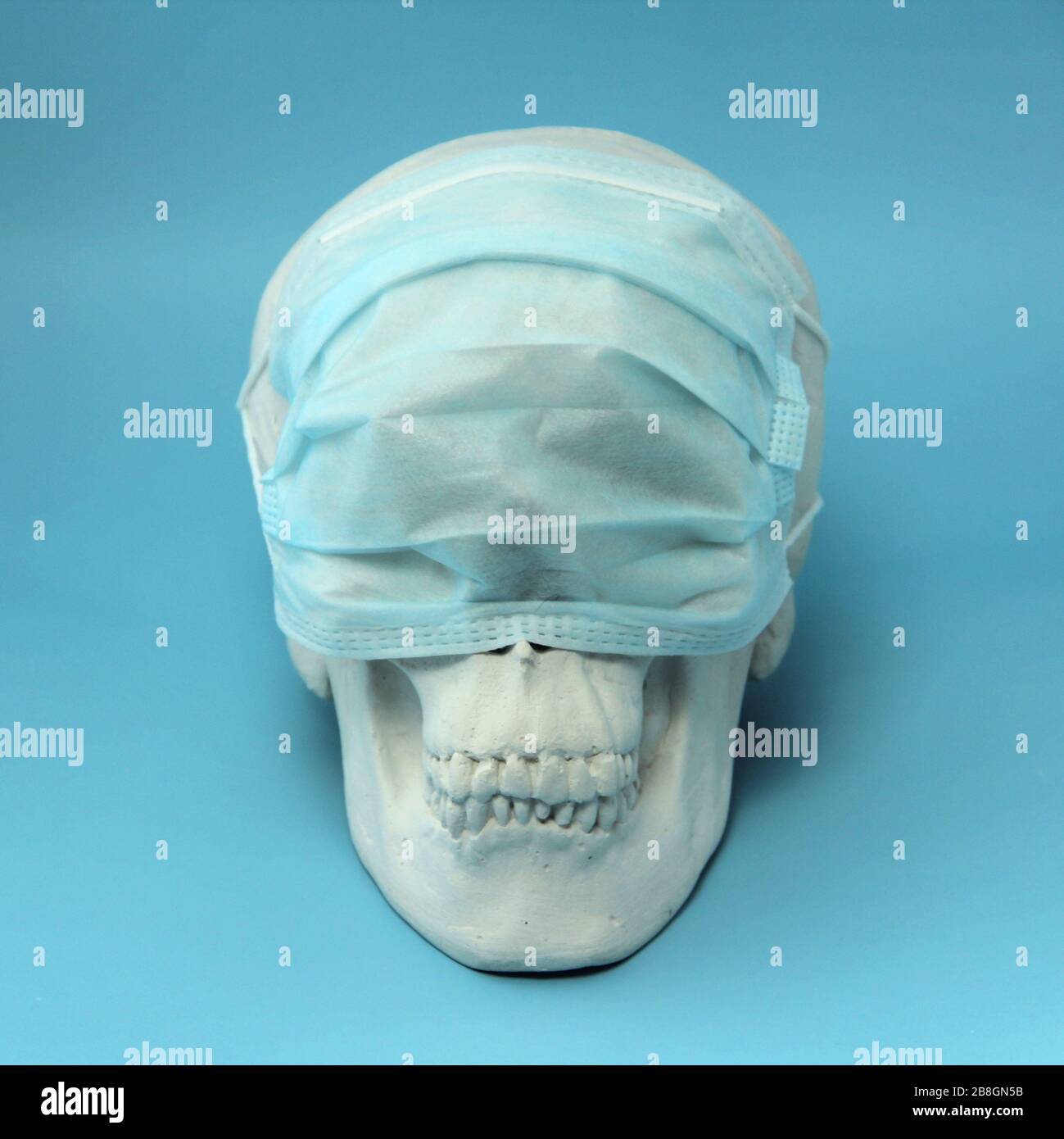 White human skull made of gypsum on a blue background with a medical mask on his eyes. The concept of health, medicine, epidemic and coronovirus. Stock Photo