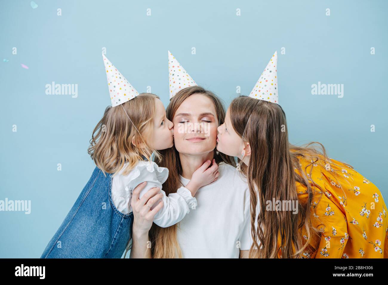 Two little girls kissing their mother on a cheek, all in party cones over blue Stock Photo