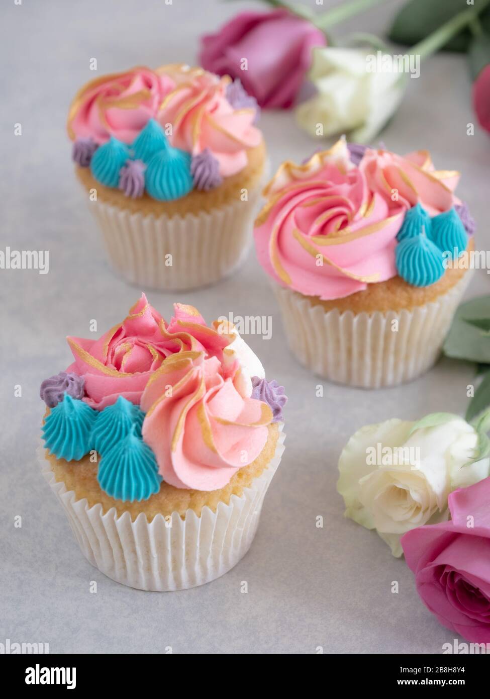 Three cupcakes with beautiful frosting among some roses on a tabletop Stock Photo